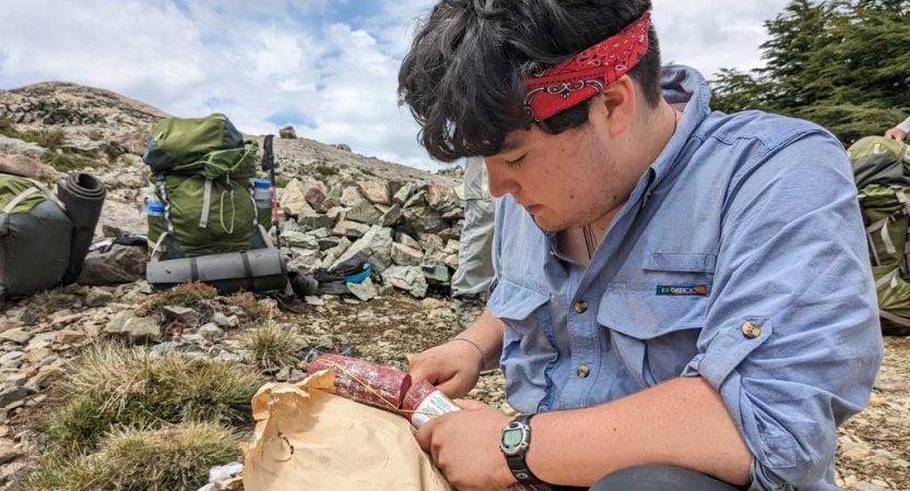 A person sits outside and cuts a piece of sausage. Backpacks are resting on the ground nearby. 